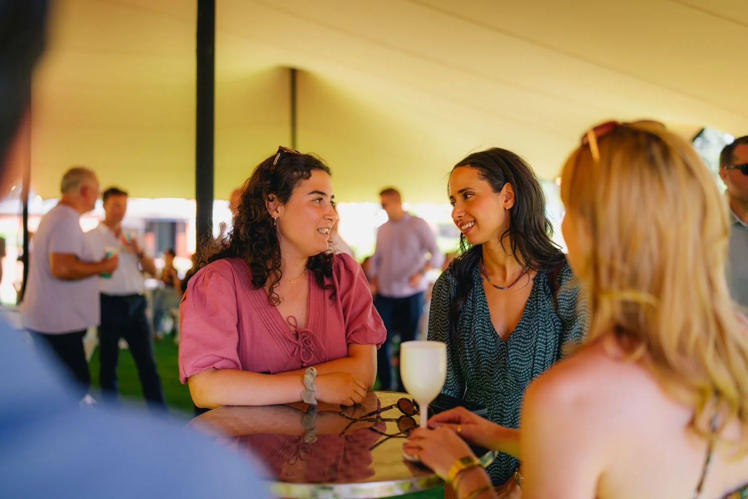 Four women are gathered around a table under a large yellow canopy at a social event. Two women in the foreground are engaged in conversation, smiling at each other. The woman on the left has curly dark hair and is wearing a pink blouse, while the wo