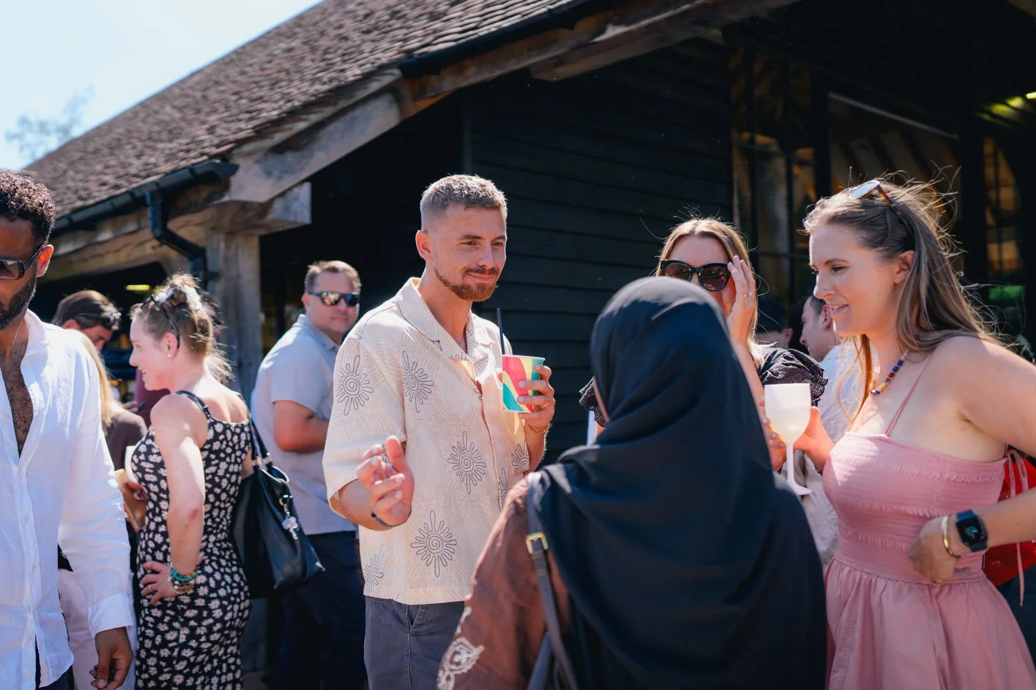 People socializing outdoors at a gathering, some holding drinks, in front of a black wooden building on a sunny day.