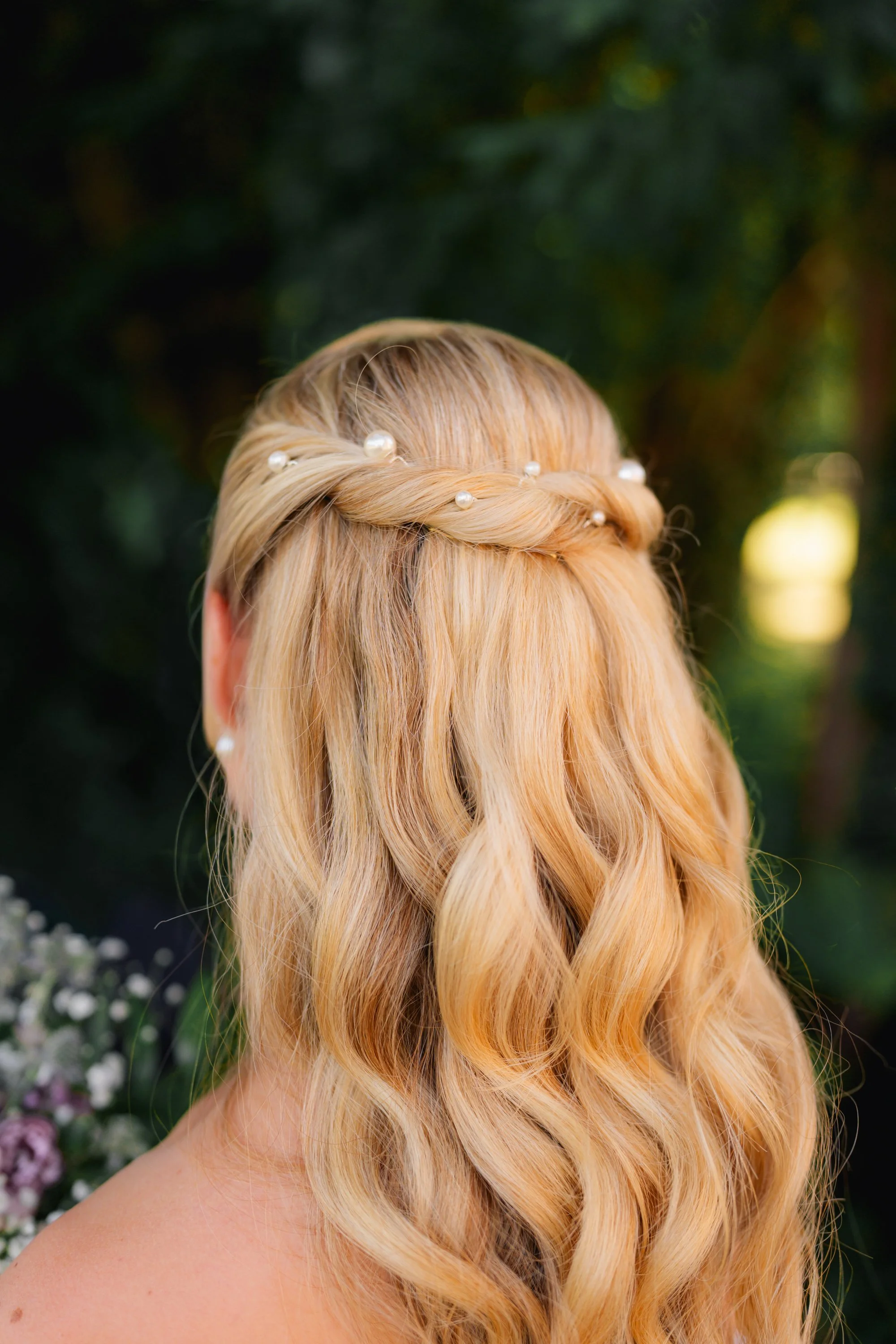 Back view of a woman with long, wavy blonde hair adorned with pearls, outdoors with blurred greenery and flowers in the background.