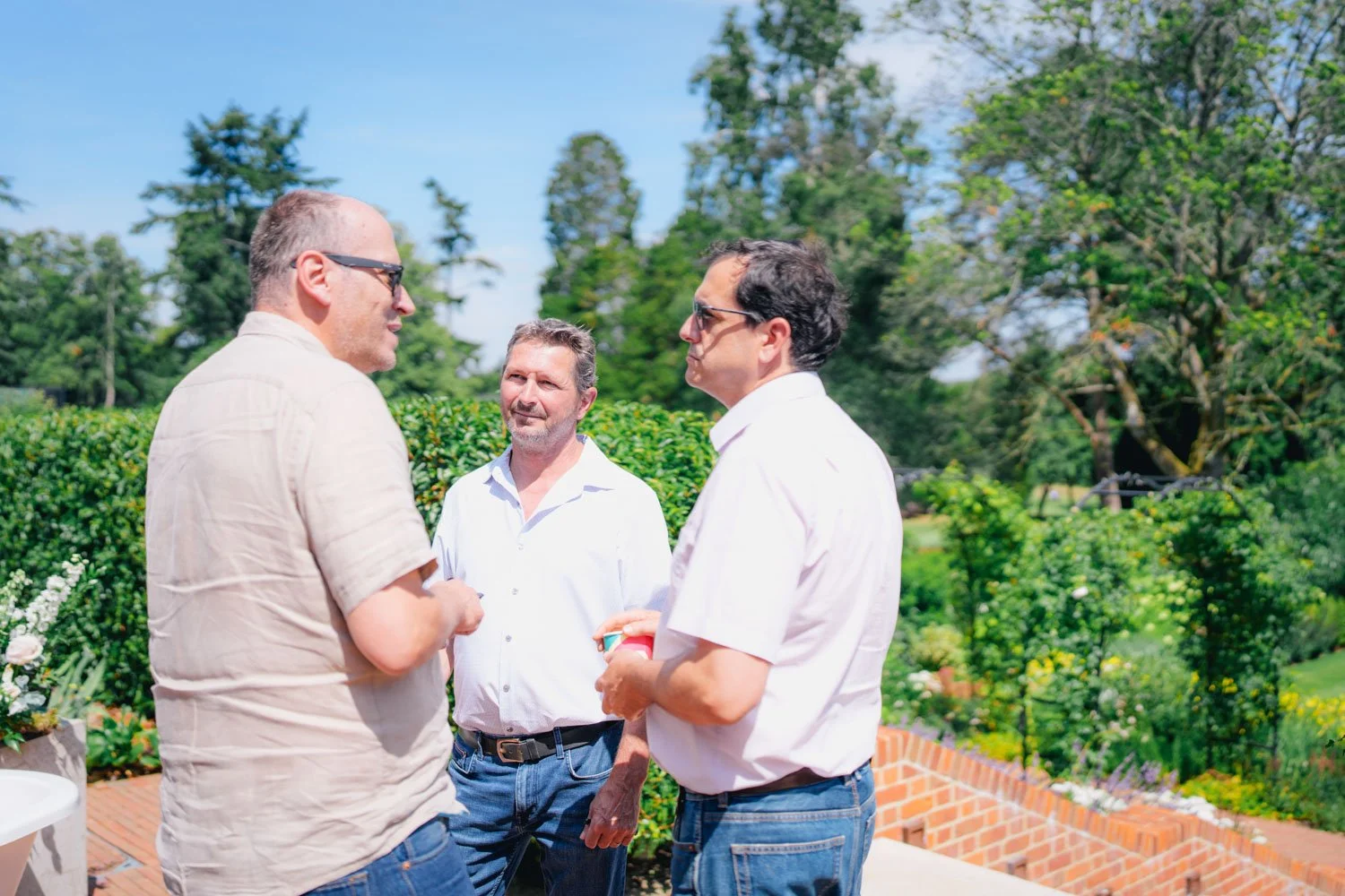 Three men conversing outdoors on a sunny day with greenery and trees in the background.