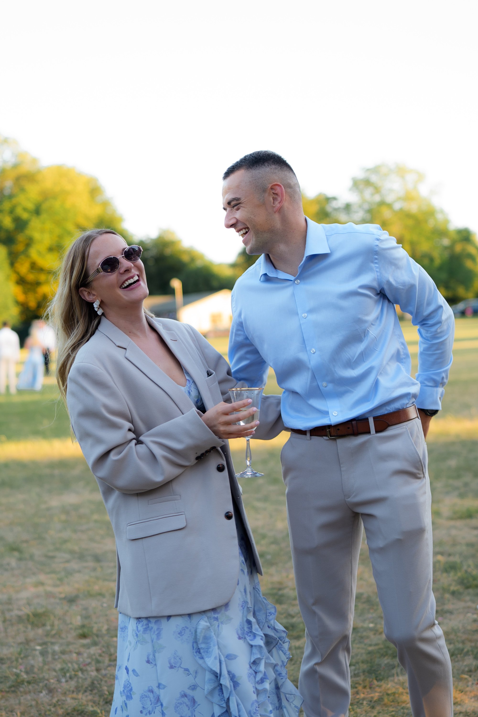 A man and woman are laughing and chatting outdoors at a social gathering in a park, holding a wine glass.