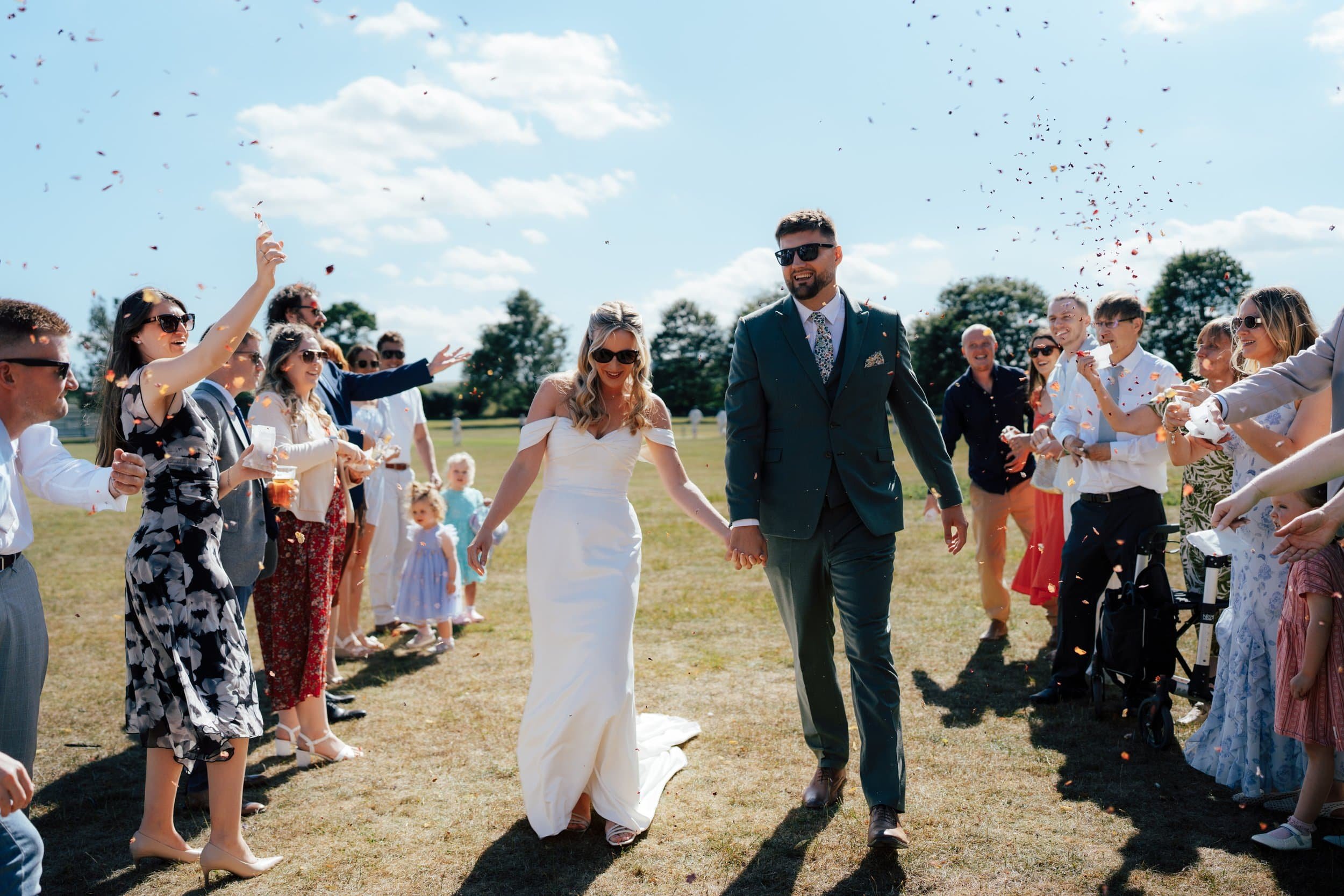 A bride and groom walking hand in hand through a crowd of wedding guests outdoors on a sunny day, with confetti in the air.