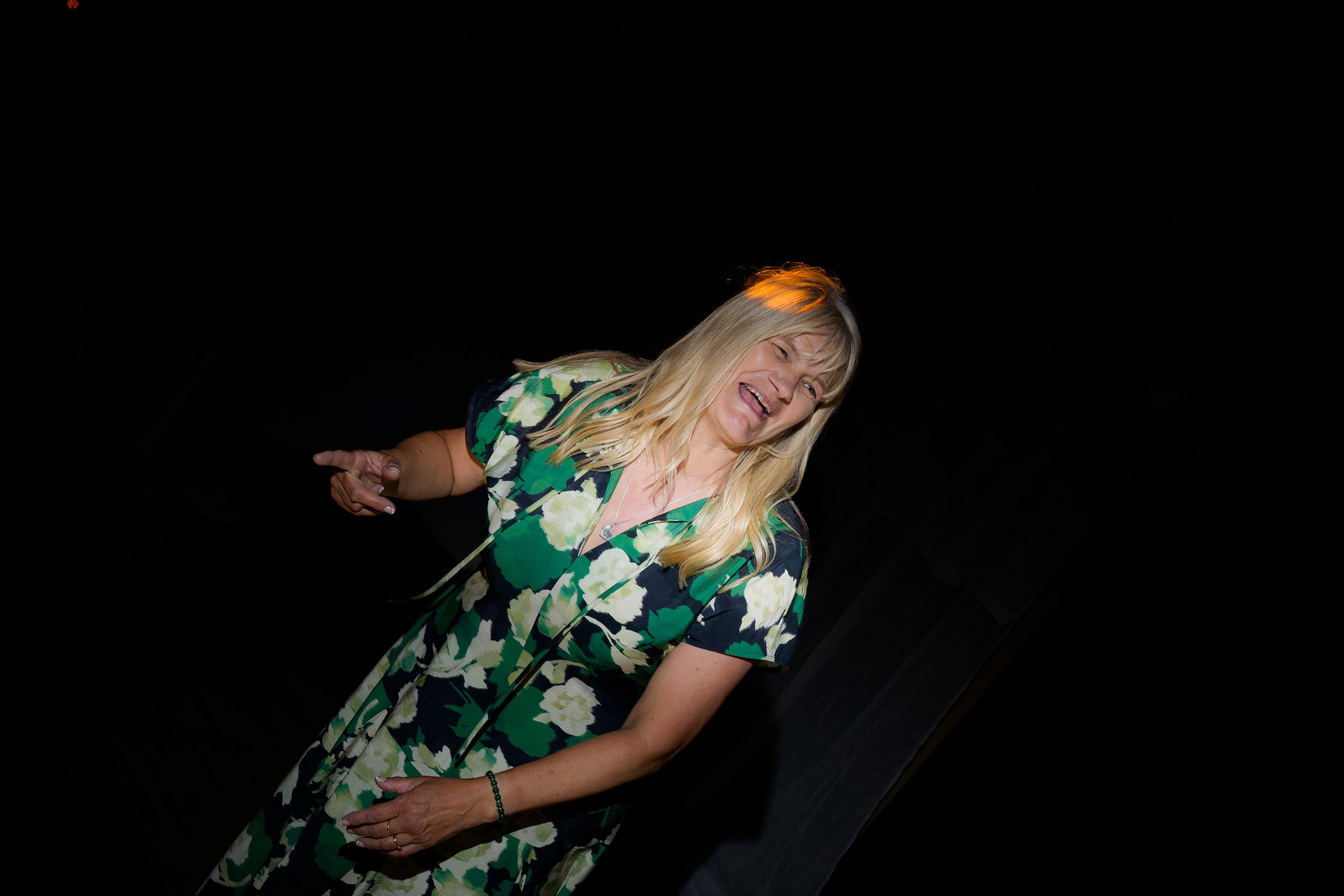 A woman with blonde hair wearing a green, white, and black floral dress, smiling and posing against a dark background.