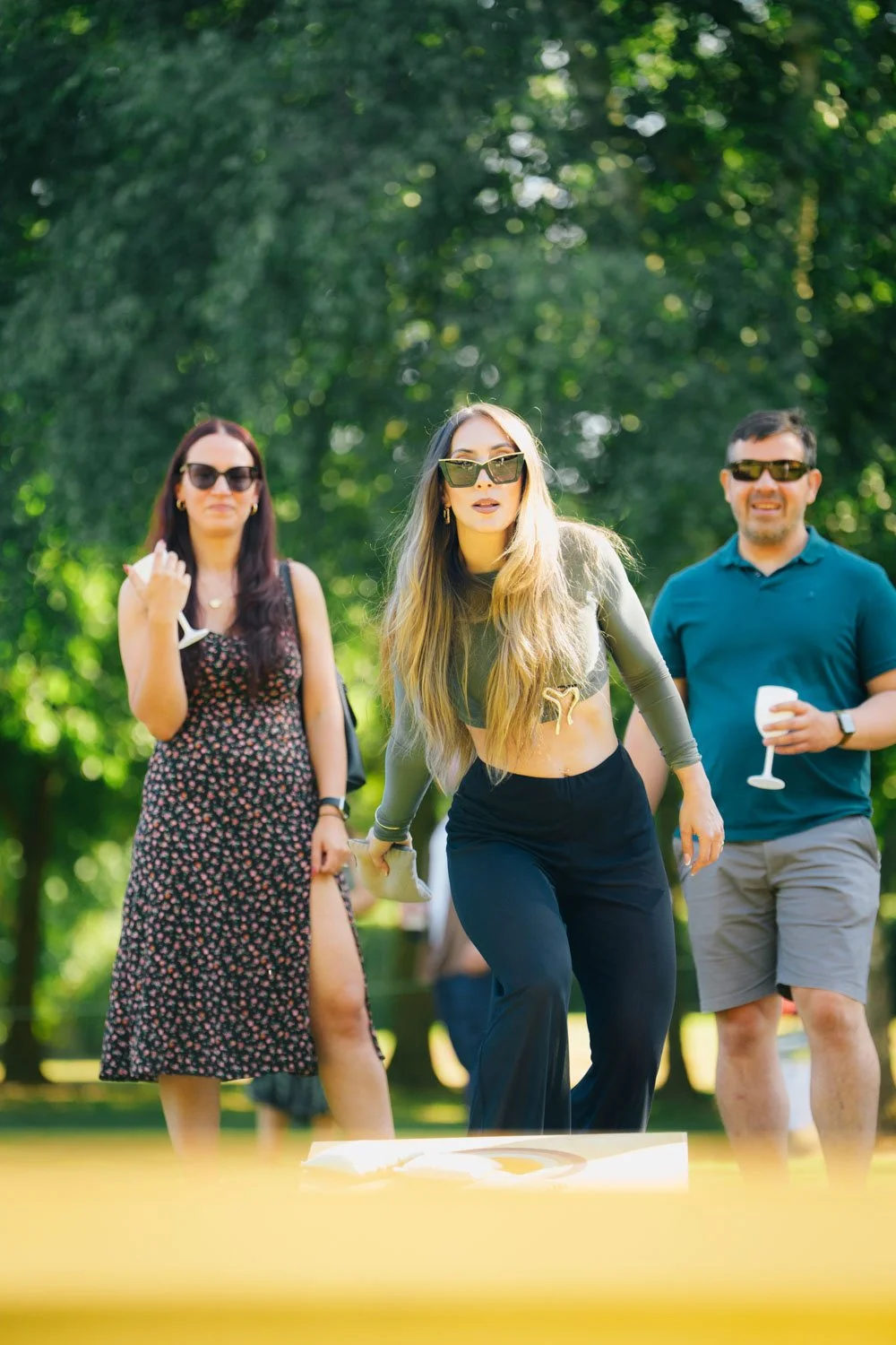 Three people outdoors, two women and one man, enjoying drinks and socializing on a sunny day with green trees in the background.
