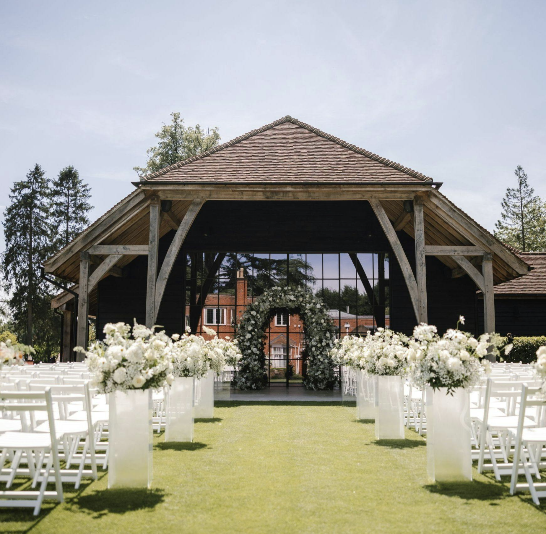 Outdoor wedding setup with rows of white chairs flanking an aisle decorated with white flowers and greenery, leading to a wooden arch with floral arrangements under a roofed structure, with trees and buildings in the background.