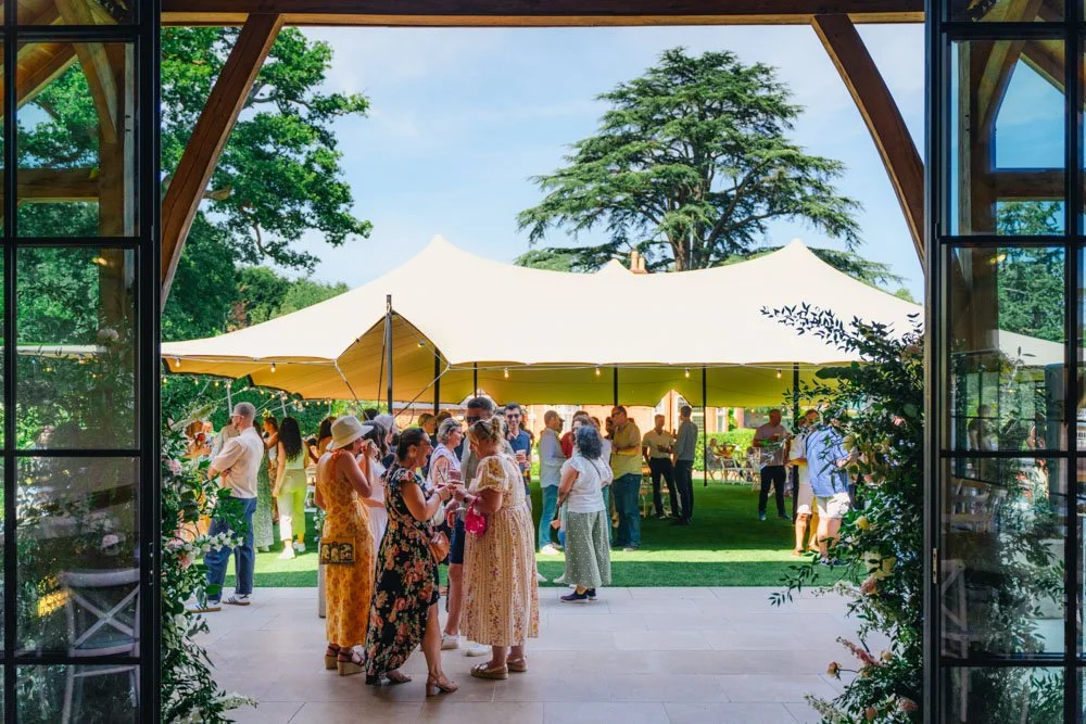 People at an outdoor gathering under a large yellow canopy tent, viewed from inside a building through an open door, with trees and a partly cloudy sky in the background.