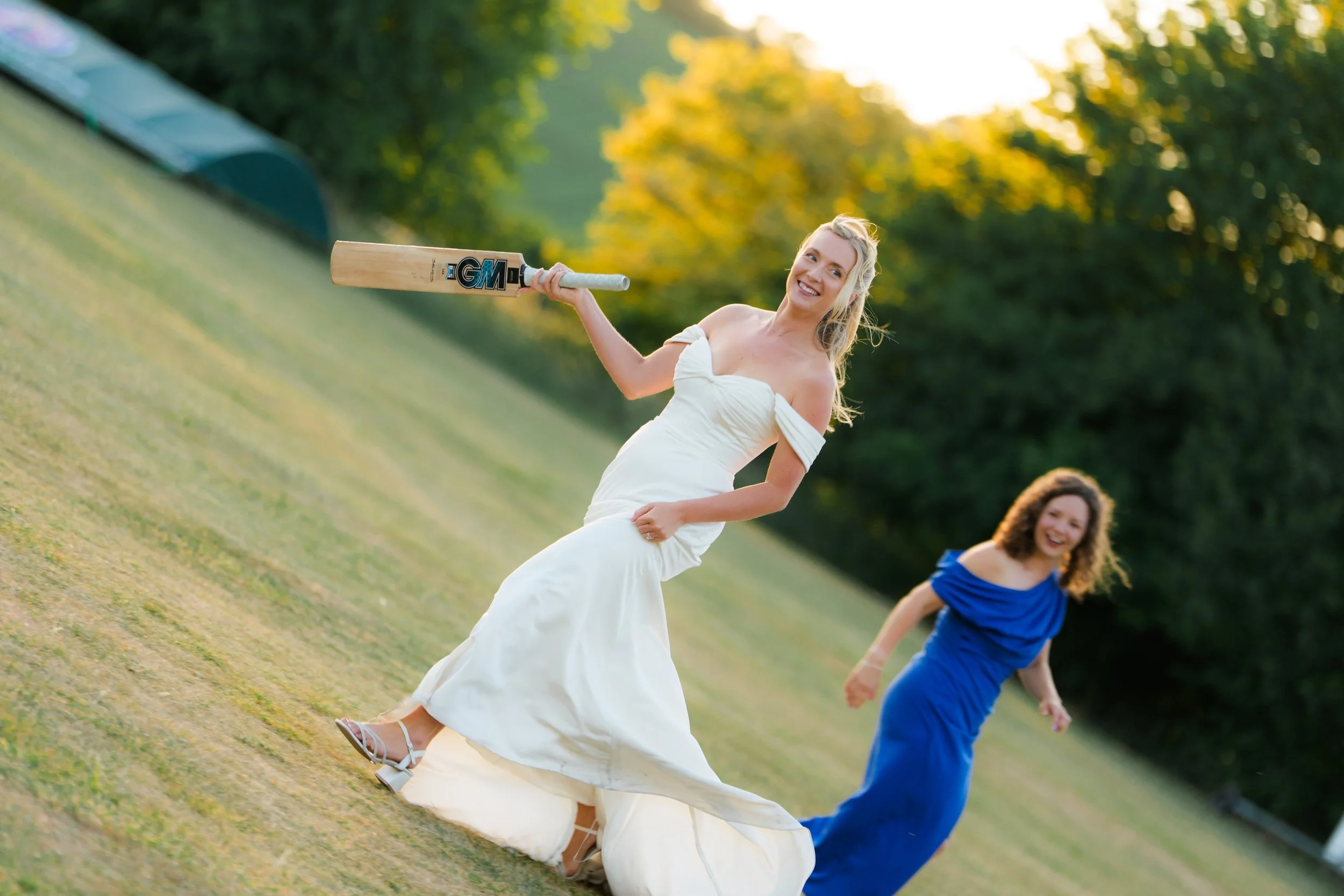 Two women wearing dresses and jumpsuits having fun outdoors in a park during sunset, one woman in a white dress holding a cricket bat, smiling. The other woman in a blue jumpsuit in the background, also smiling.
