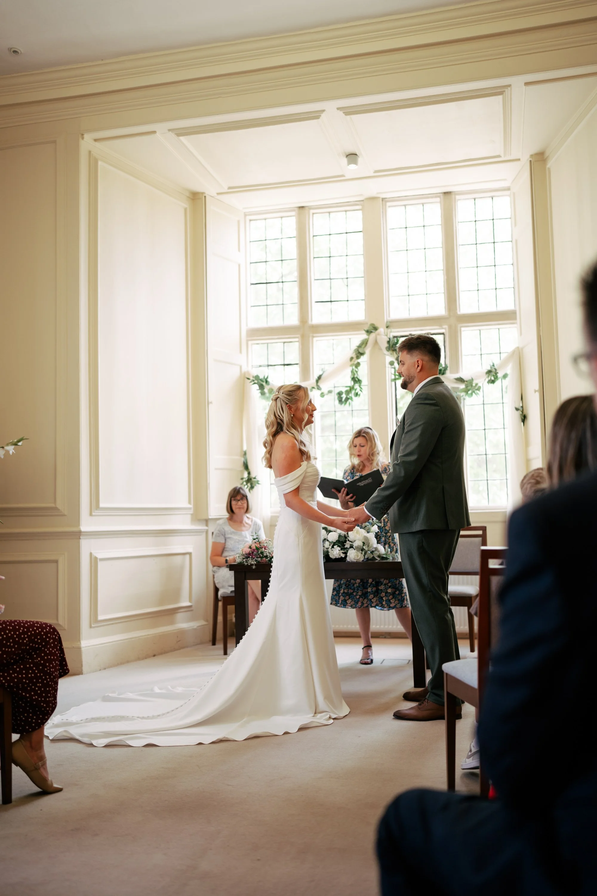 A wedding ceremony taking place inside a bright room with large windows. The bride and groom are holding hands and facing each other, with officiants and guests in the background.