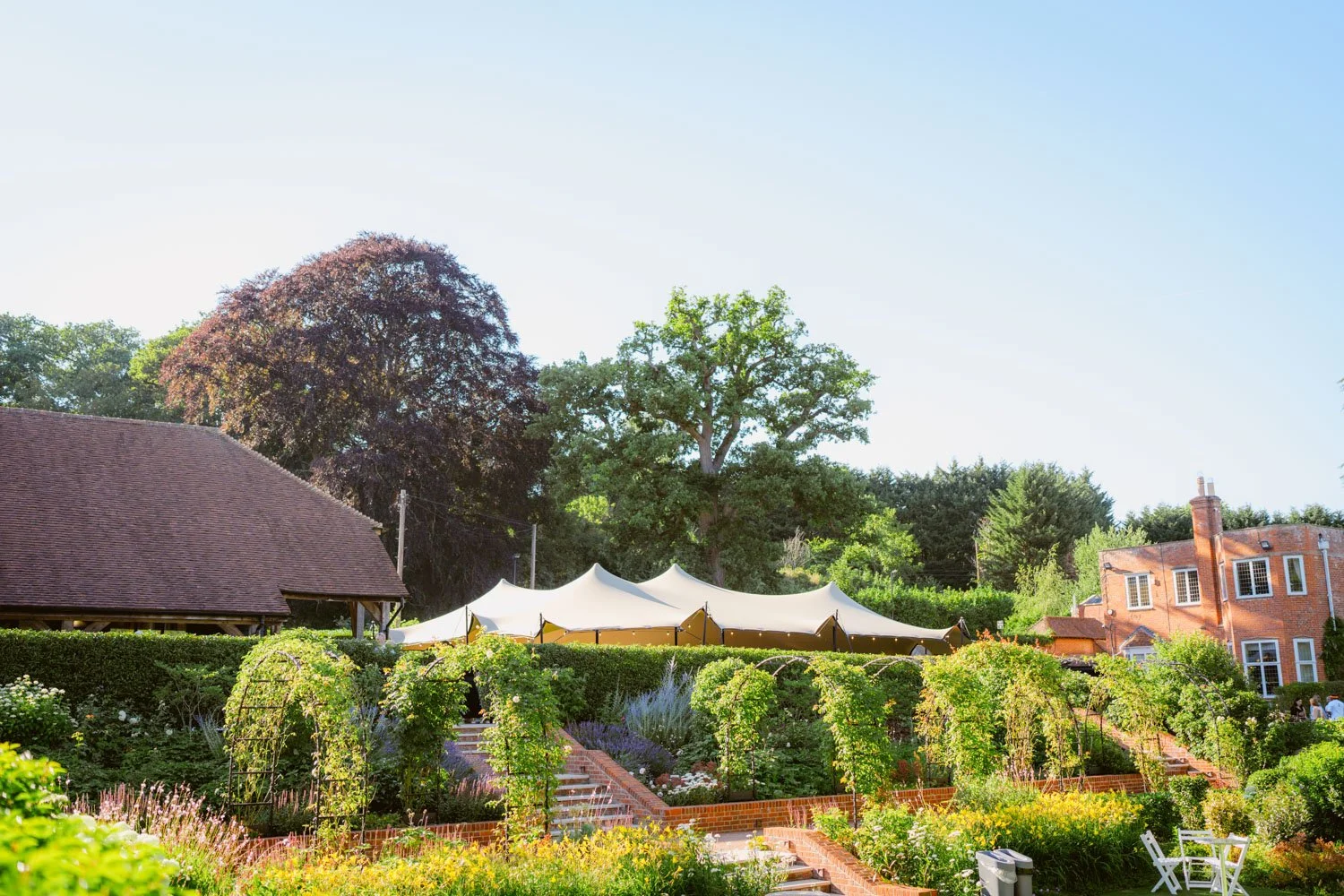 A lush garden with flower beds, trellises, and a tent canopy in a backyard with a large house and trees in the background.