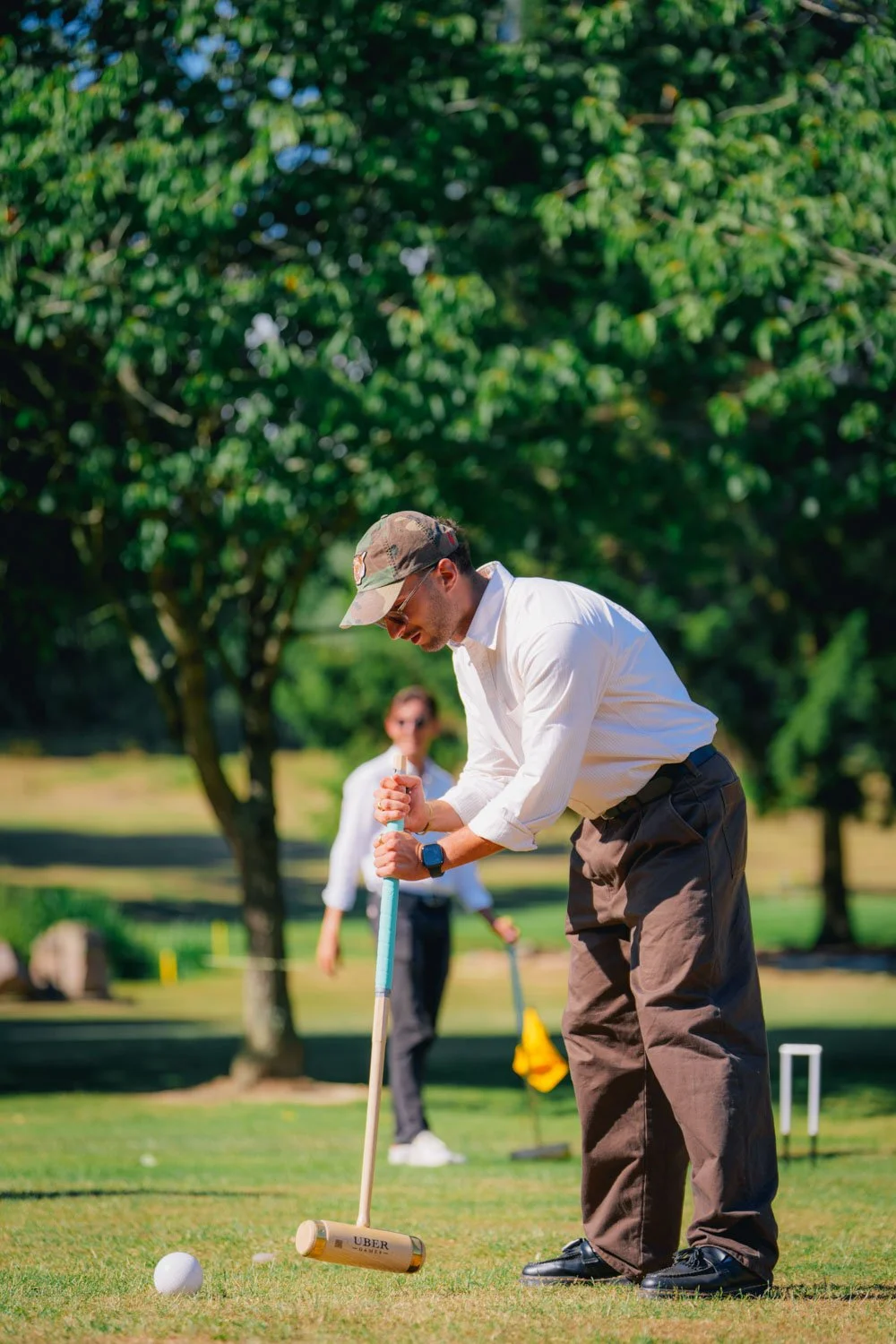 A man wearing a white shirt, cargo pants, and a camo hat is playing mini-golf outdoors. He is holding a mallet and aiming at a golf ball. A woman in the background is watching and holding a golf club, with trees and a bright, sunny sky around them.