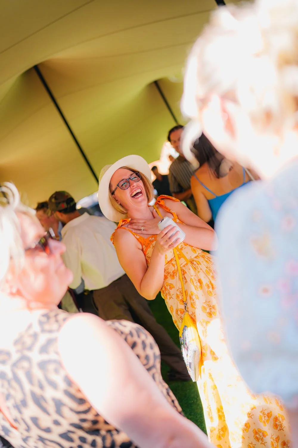 A woman in a yellow floral dress and white sunhat laughing and holding a drink at a social event with others around her under a yellow canopy.
