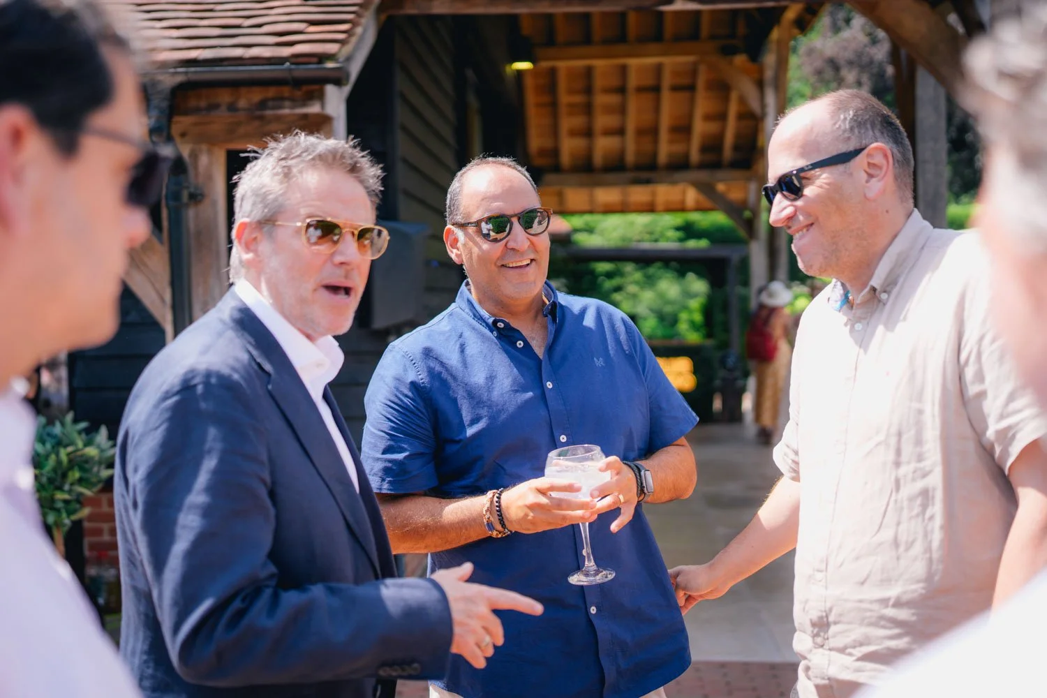 Group of casually dressed men wearing sunglasses engaged in conversation outdoors, with a rustic wooden building in the background.