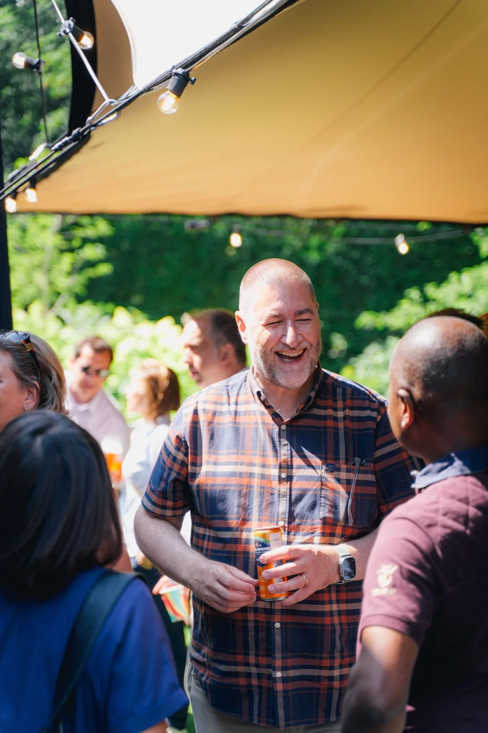 People socializing outdoors under string lights and a canopy, with trees in the background. A man in a plaid shirt is smiling and holding a drink while talking to another person.