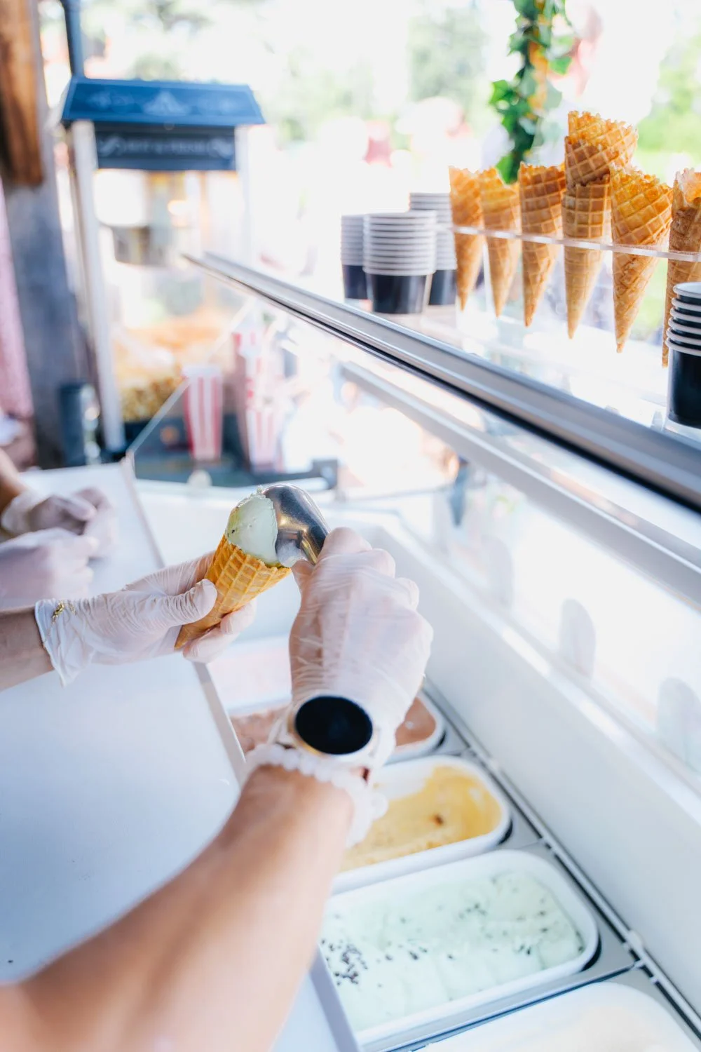 Person scooping ice cream into a cone at an ice cream shop with cups and waffle cones displayed behind the counter.