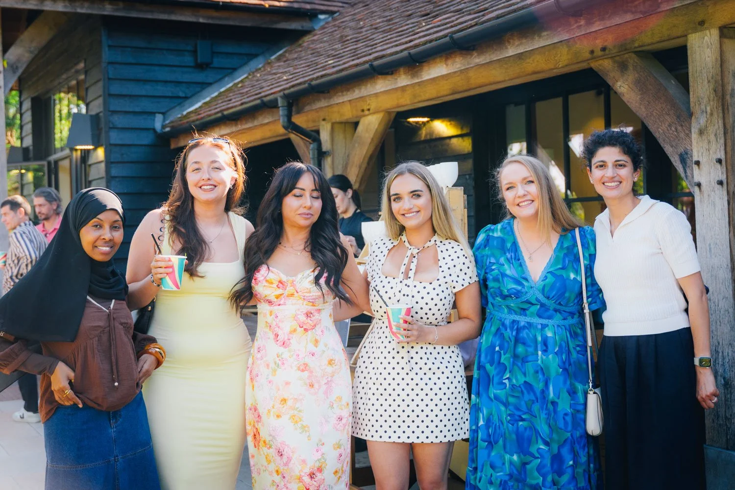 Group of six women standing outdoors in front of a building, smiling at the camera, some holding colorful cups.