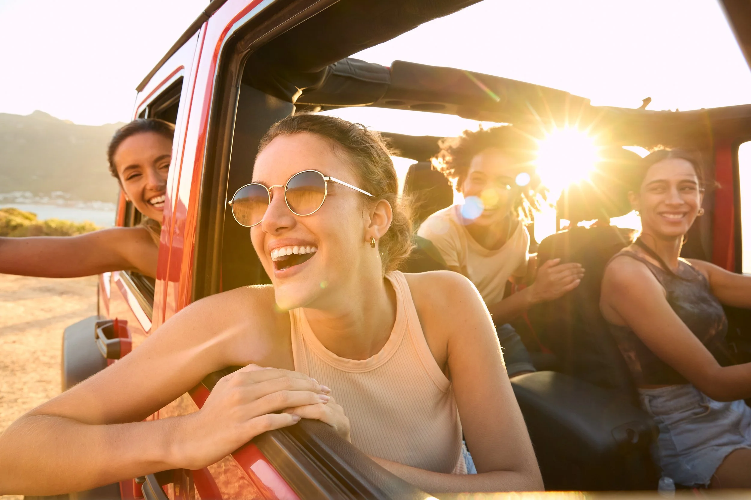 A group of five friends enjoying a road trip in a red jeep on a sunny day, smiling and having fun.