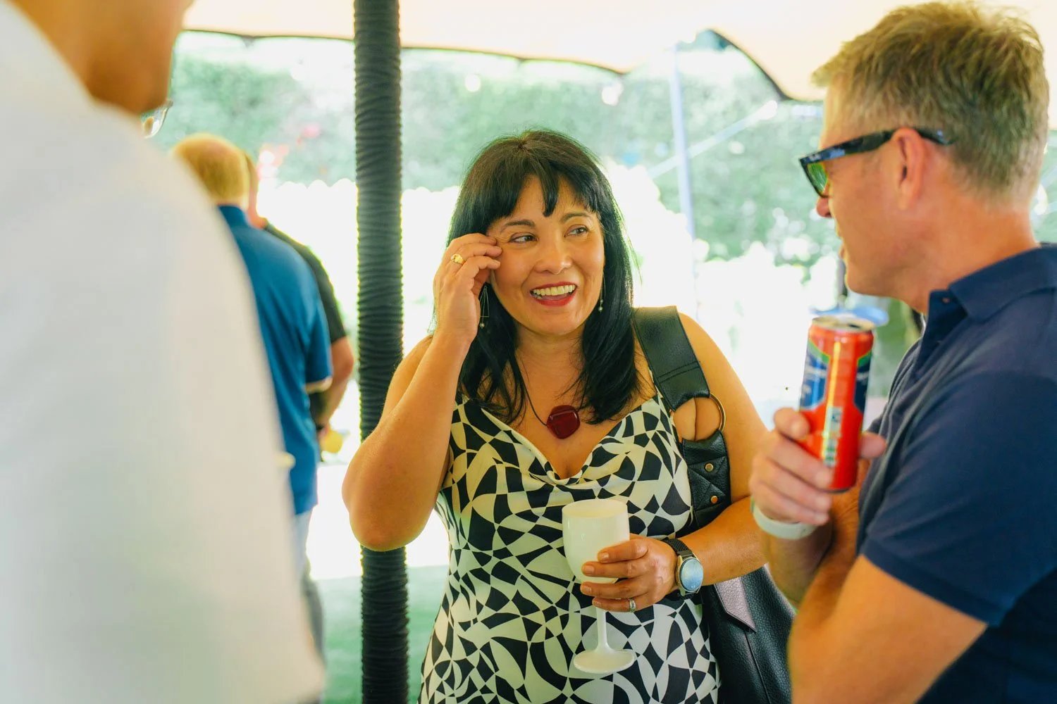 A woman with black hair and a patterned dress is smiling and talking to a man holding a soda can. She is holding a white cup and wearing a necklace with a large red pendant, with a black shoulder bag. The setting appears to be a social gathering outd