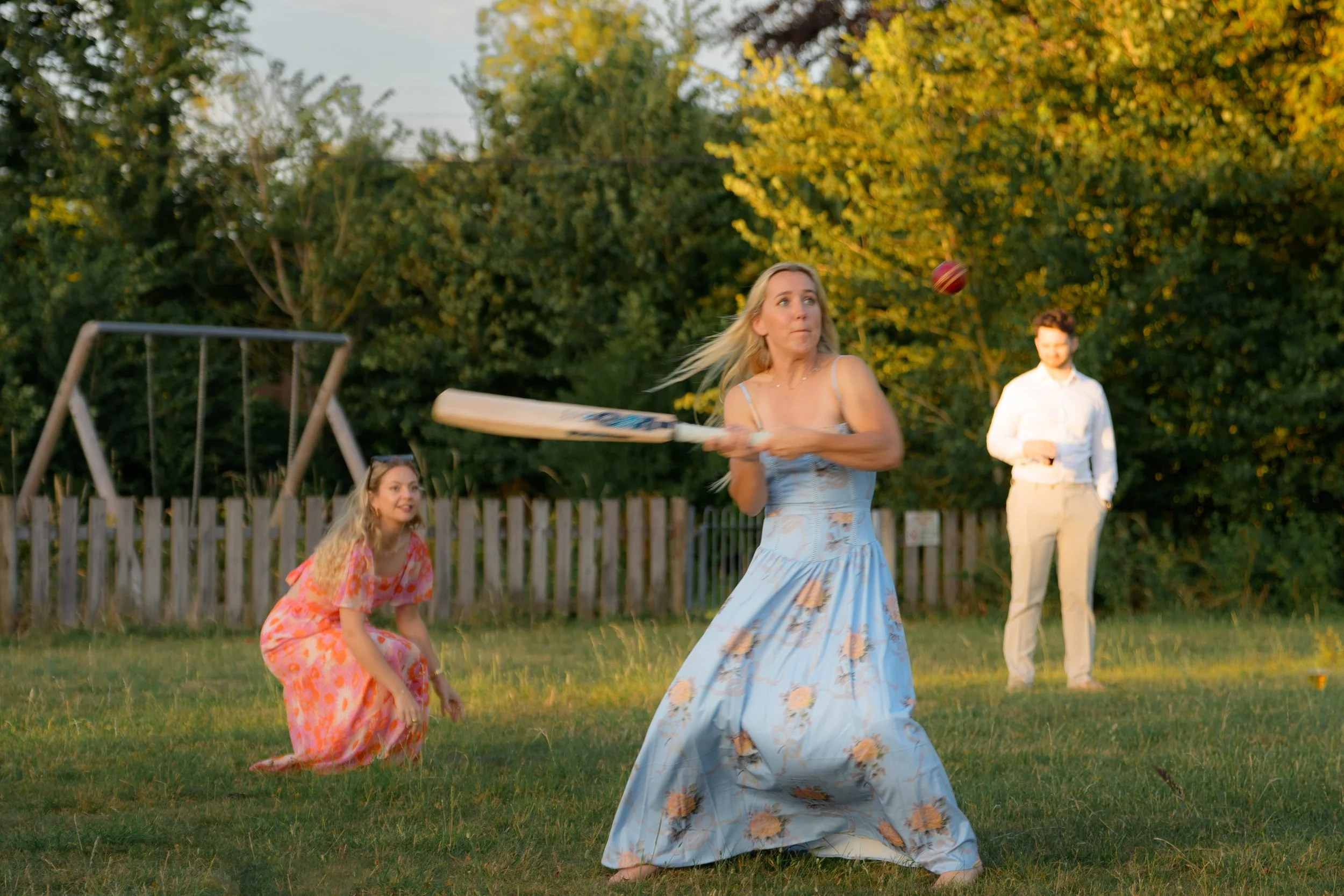 A woman in a blue floral dress is playing cricket with a red cricket ball in a backyard, with a girl in a pink dress crouching near the grass and a man in white standing in the background.
