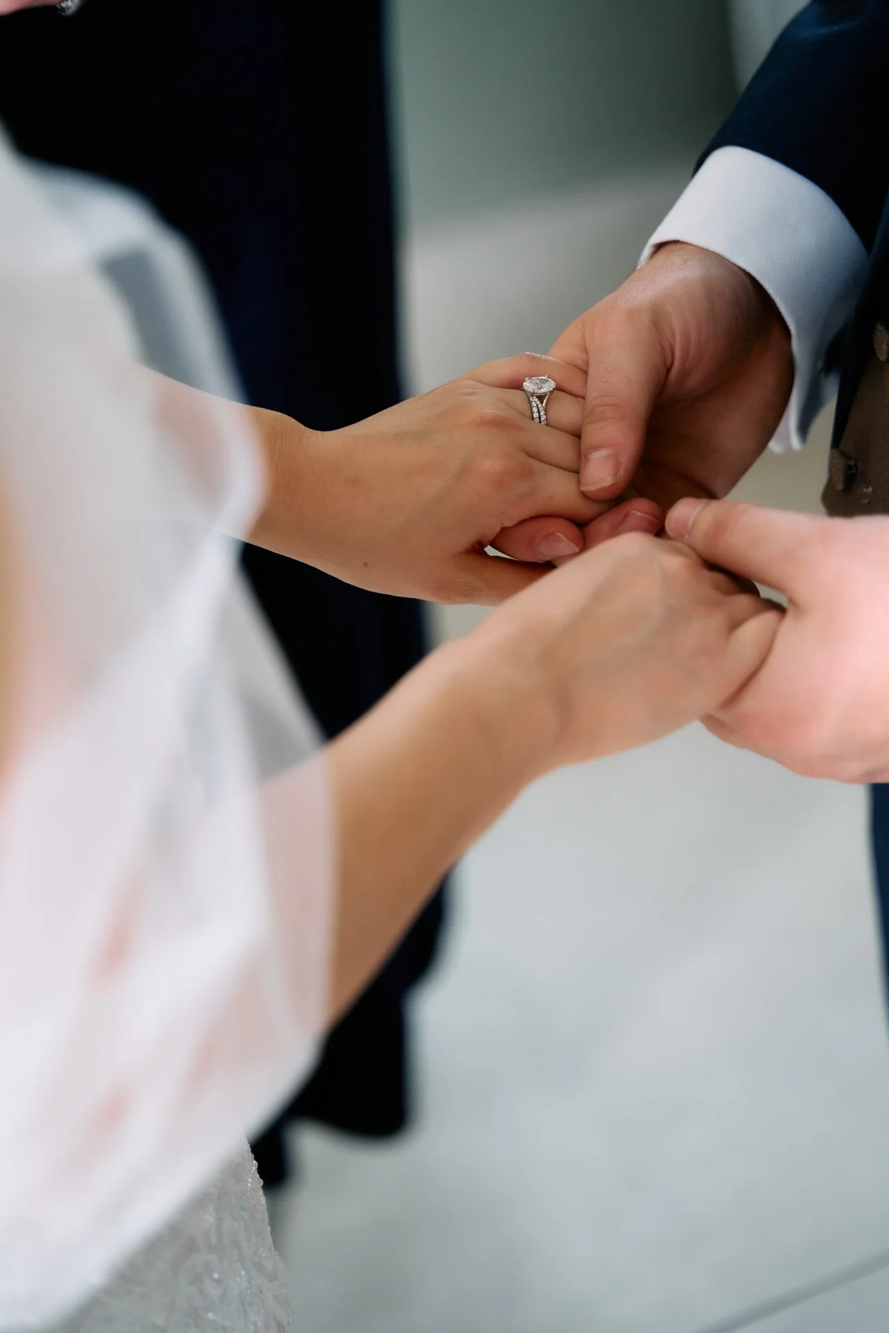 Close-up of a bride and groom holding hands during a wedding ceremony, with the groom wearing a suit and the bride wearing a wedding dress, showing her engagement ring and wedding band.
