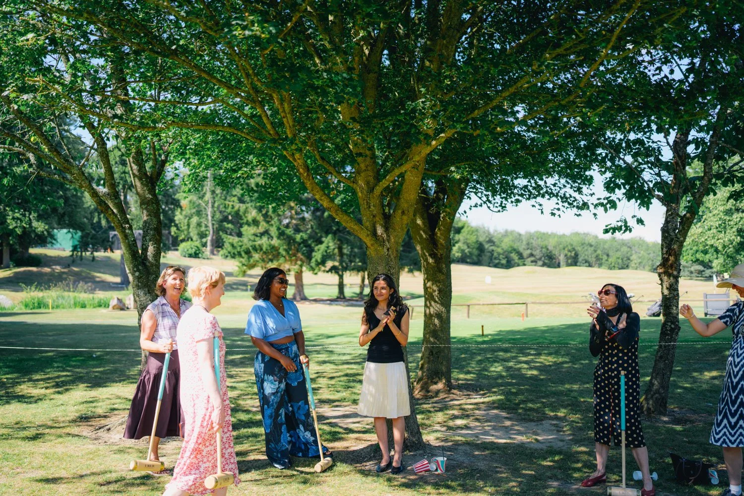 A group of women playing a game of croquet outdoors in a park with green grass, trees, and clear skies.