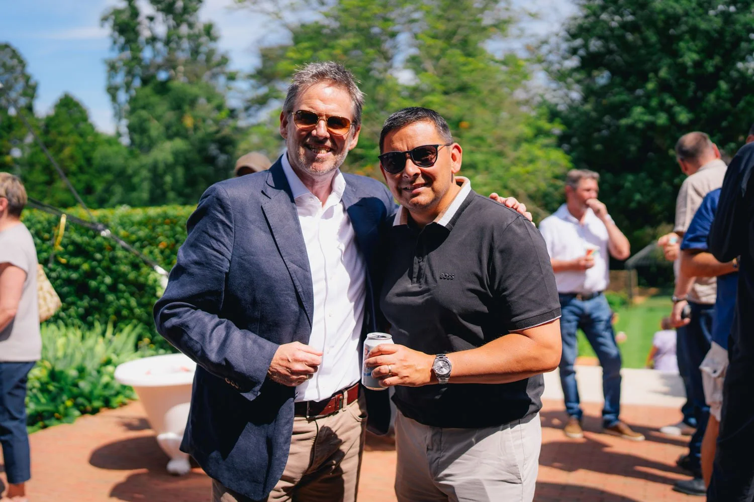Two men with sunglasses smiling at an outdoor social gathering, each holding a drink, with people and greenery in the background on a sunny day.