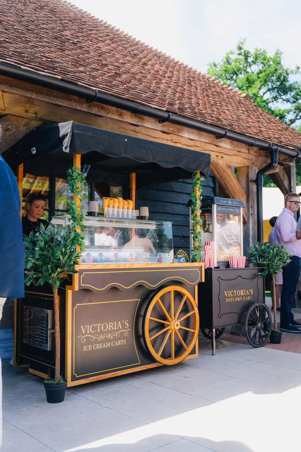 A vintage-style ice cream cart with black and yellow colors, labeled 'Victoria's Ice Cream Carts,' parked outside a wooden building with a red tiled roof. The cart has a glass display case with ice cream cones and small cups, some greenery decoration