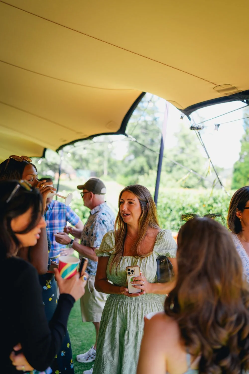 A group of people at an outdoor gathering under a canopy or tent, with green trees in the background, chatting and holding drinks and phones.