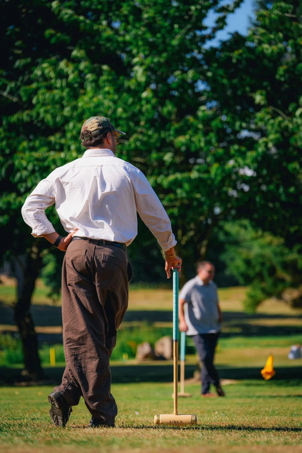 A man standing on a lush golf course, holding a wooden mallet, preparing for a game, with trees and another person in the background on a sunny day.