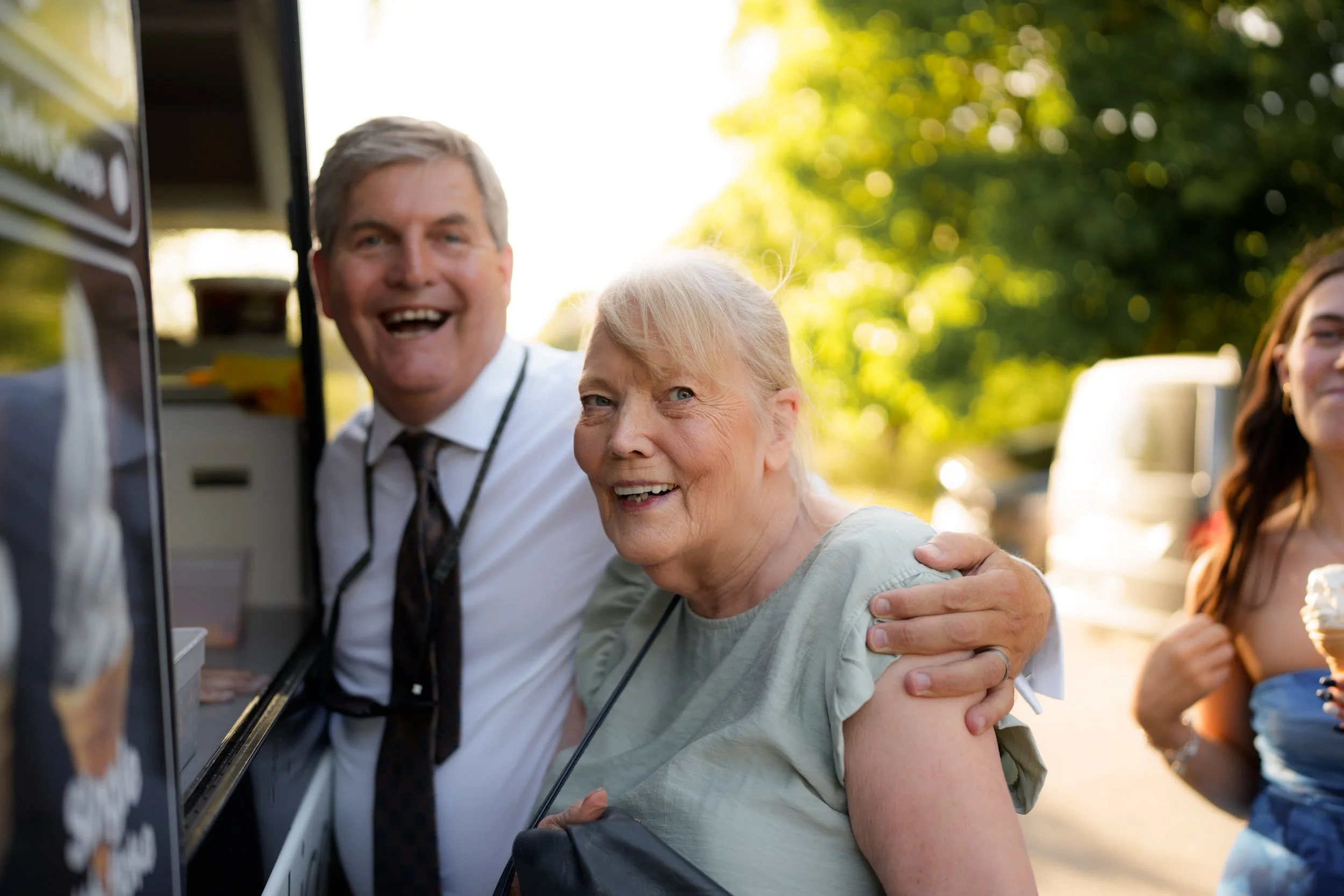 An elderly woman with blonde hair and a man in a white shirt smiling and hugging at an outdoor event, with trees and people in the background.