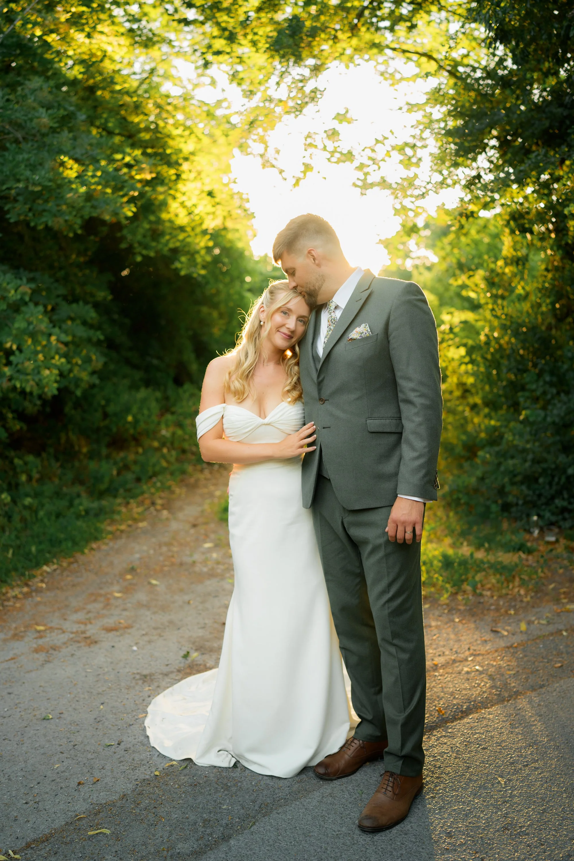 A bride in a white strapless wedding gown and a groom in a gray suit with a floral tie, standing on a trail surrounded by green trees, with sunlight glowing behind them.