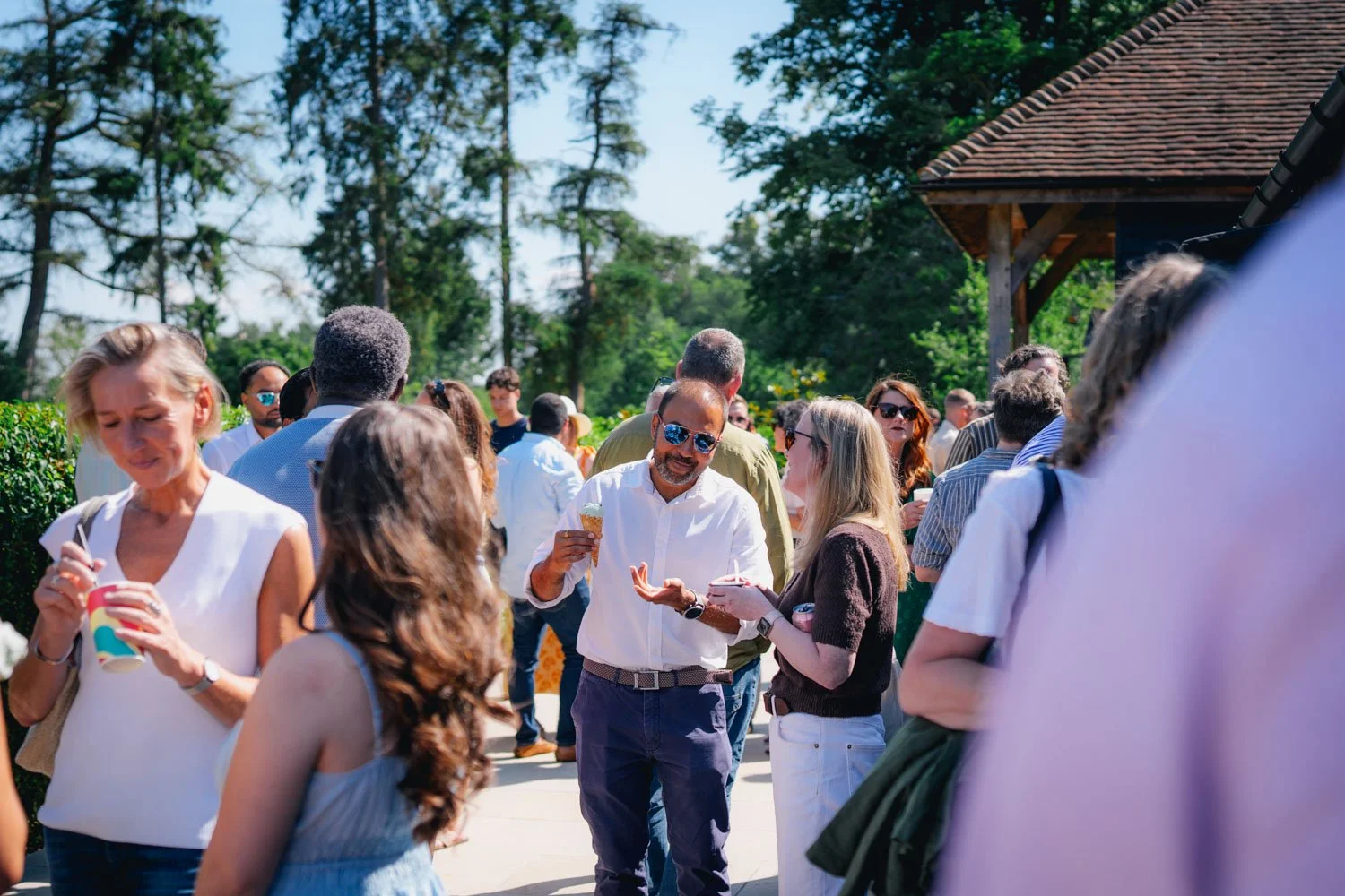 People socializing outdoors on a sunny day, some holding ice cream or drinks, in a green area with trees and a wooden building.