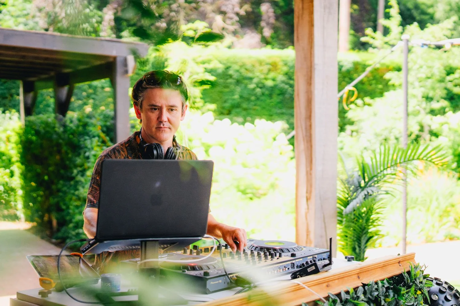 A male DJ wearing headphones working at an outdoor setup with a laptop and a DJ controller, surrounded by lush green foliage and trees.