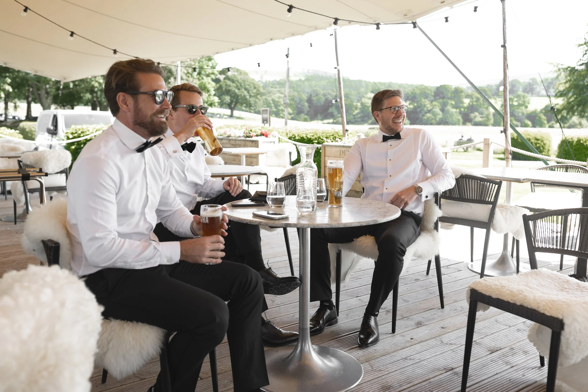 Four men in tuxedos and bow ties sitting at a round table on a patio, enjoying drinks and smiling, with a scenic outdoor background.