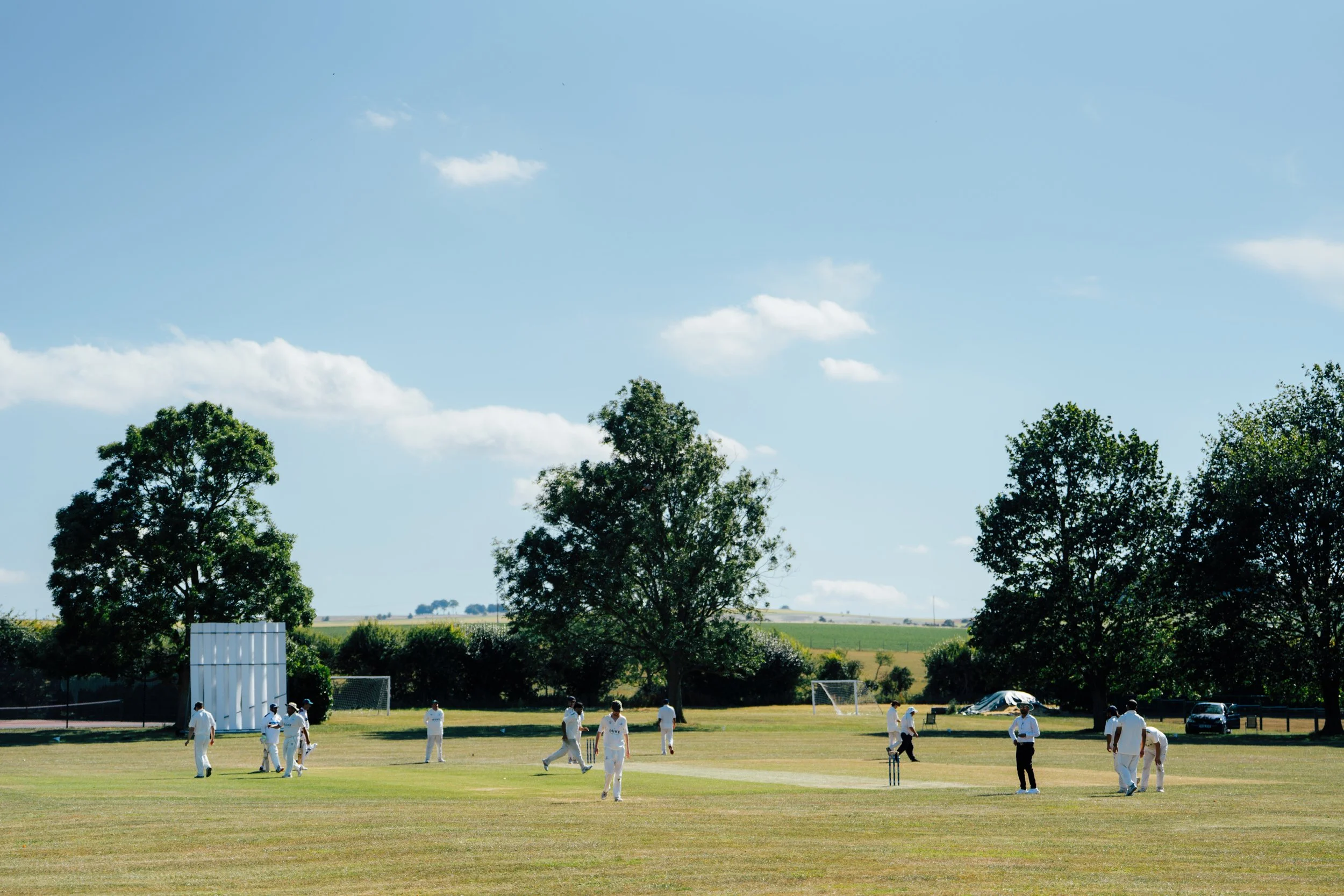 Cricket players on a field with spectators and trees in the background under a blue sky with some clouds.