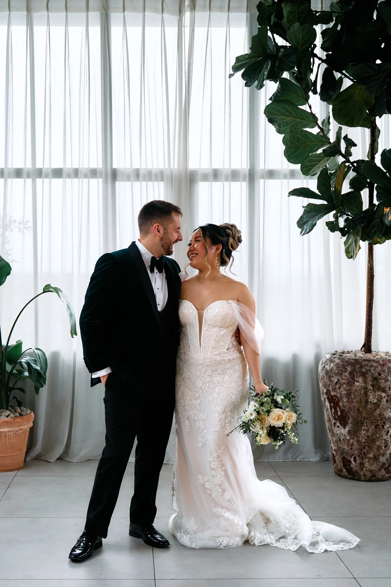 A bride and groom standing close together indoors, smiling at each other, with large potted plants and white curtains in the background.