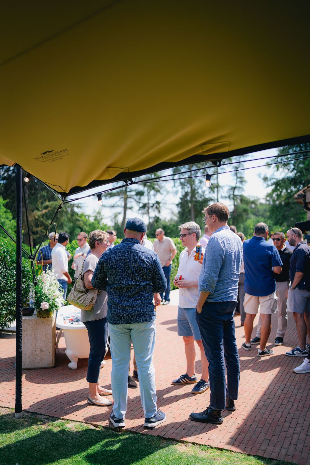 Group of people socializing outdoors under a canopy, some holding drinks, in a garden setting on a sunny day.