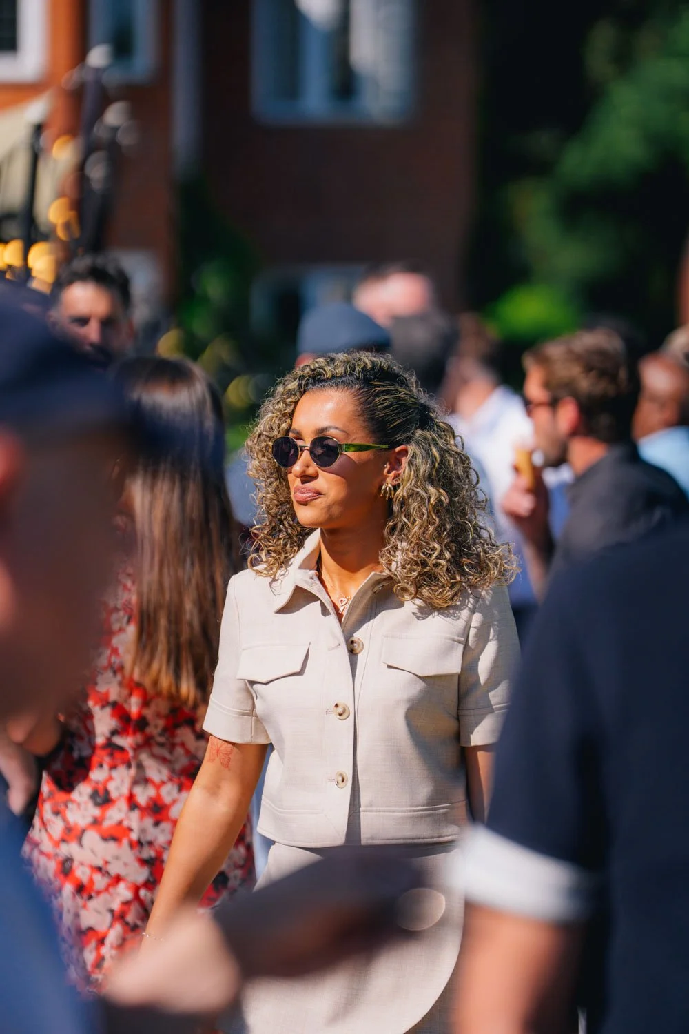 A woman with curly hair and sunglasses standing at an outdoor social gathering, surrounded by people.