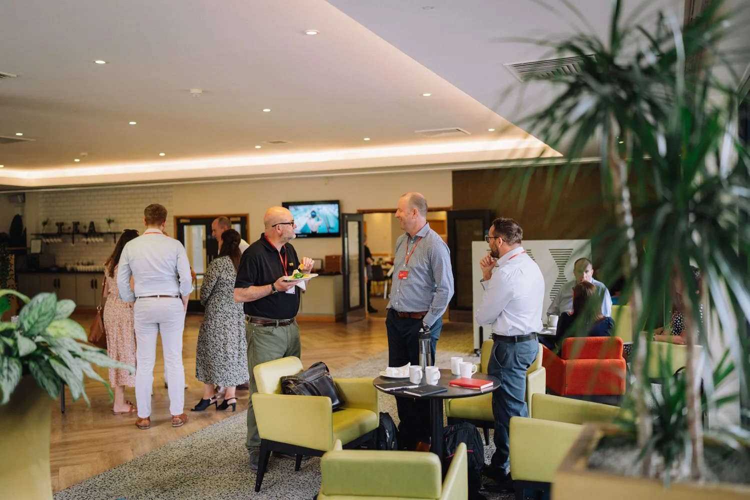 People networking and conversing in a modern hotel lounge, with some seated and some standing, in a well-lit room with a TV on the wall and a refreshment area in the background.