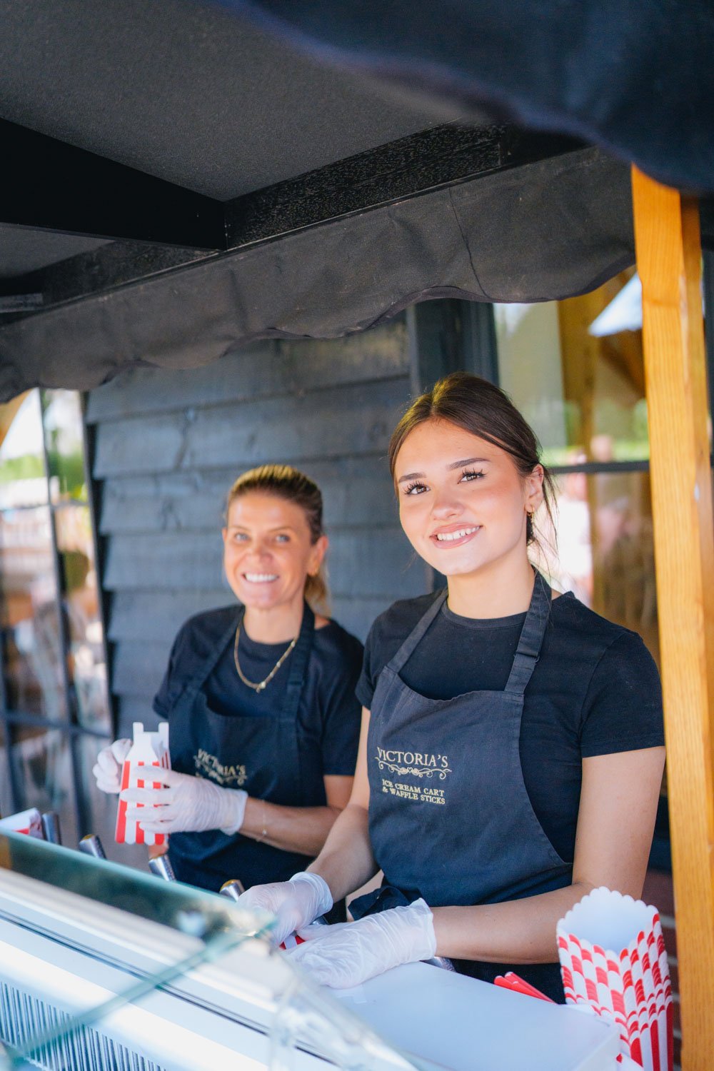 Two women working at a wood ice cream cart, smiling, wearing black aprons with 'Victoria's' logo, and white gloves.