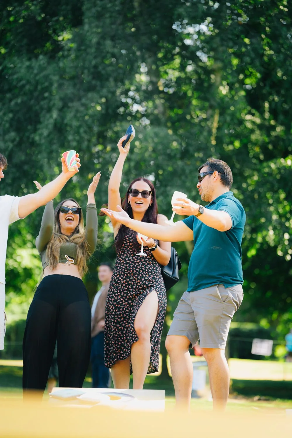 A group of people celebrating outdoors with drinks and dancing in a park with green trees.