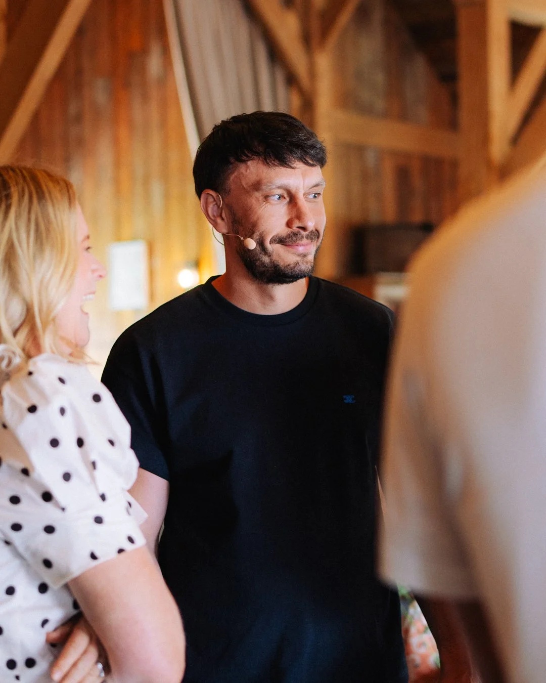 A man with dark hair and a beard wearing a black t-shirt, smiling, with a woman with blonde hair and a polka dot dress beside him, in a wooden barn setting.