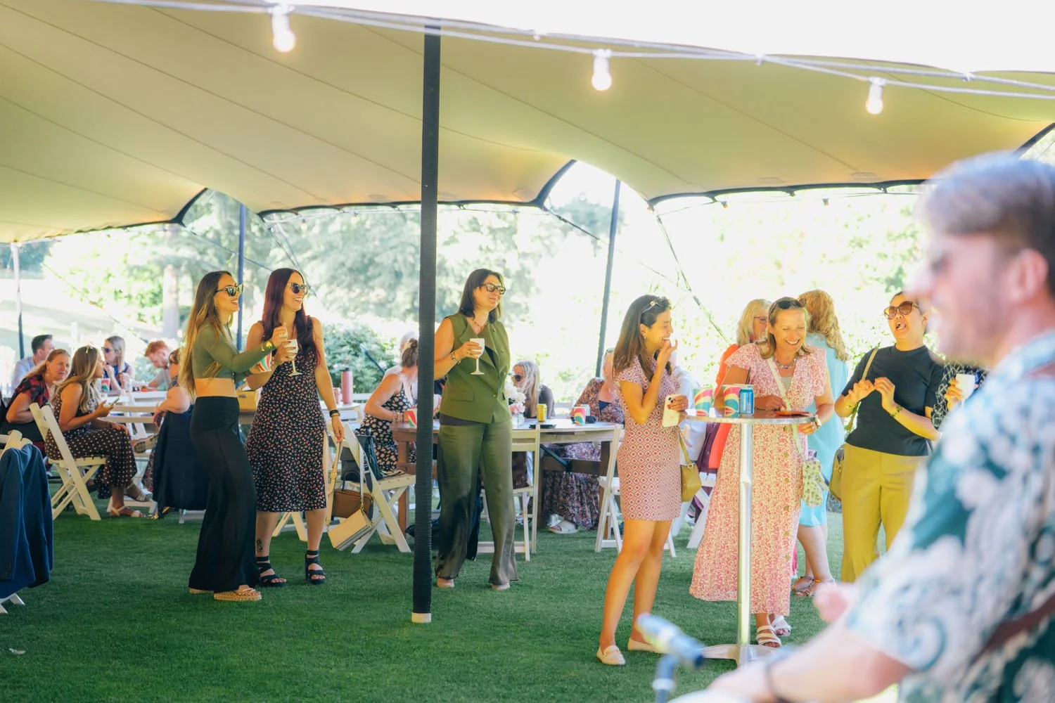 People socializing under a tent at an outdoor gathering, some holding drinks and smiling.