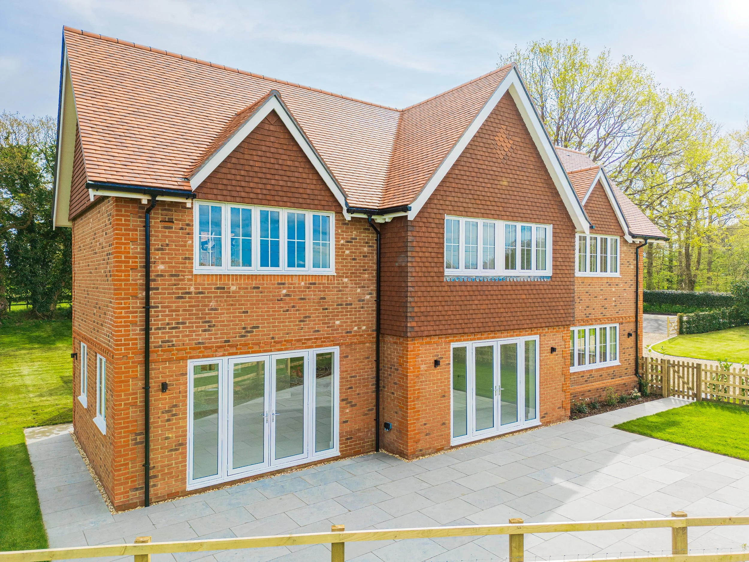 A large brick house with a red tiled roof, multiple windows, and a paved backyard with a grassy area and wooden fence.