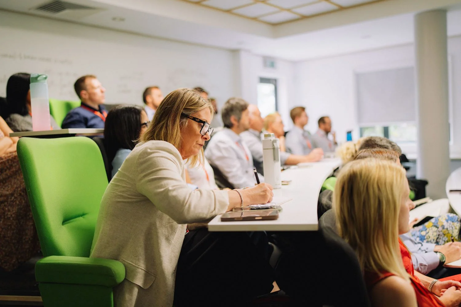 People attending a conference or seminar in a classroom-style setting with green chairs and a whiteboard.
