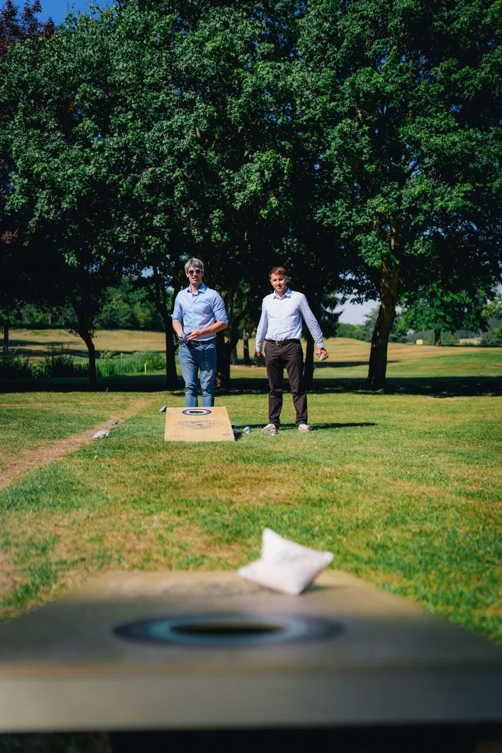 Two men playing cornhole on a grassy field with trees in the background, one is wearing sunglasses and the other is holding a bean bag.