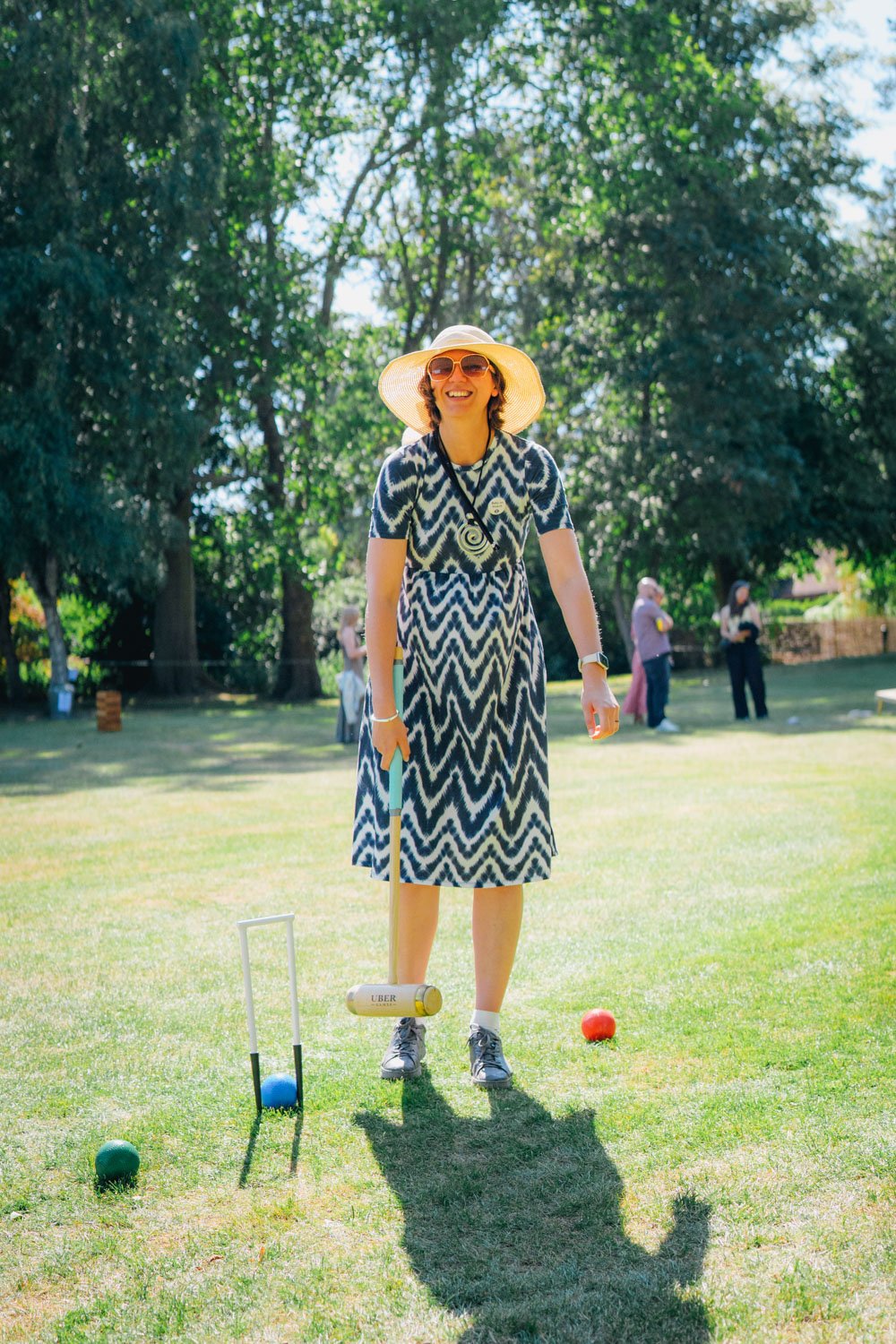 Woman smiling on a grassy field during outdoor game, wearing summer hat, sunglasses, a patterned dress, and sneakers, with bocce balls and a launcher nearby.