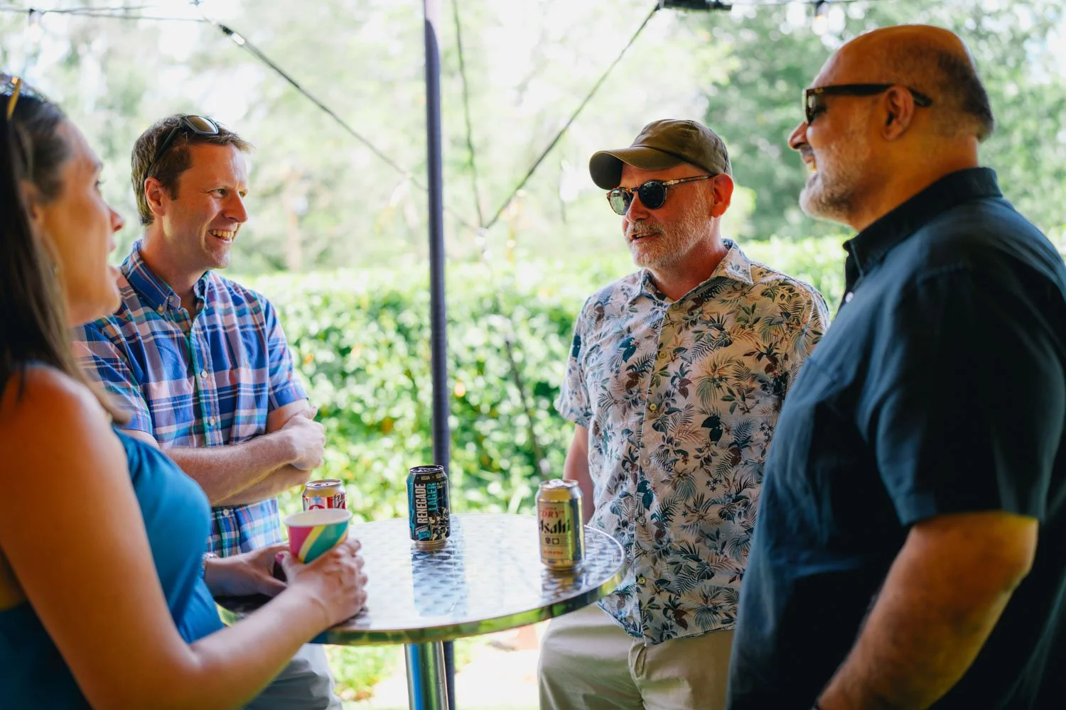 Four adults standing around a high-top outdoor table, chatting and smiling, with beverages on the table, in a garden setting.