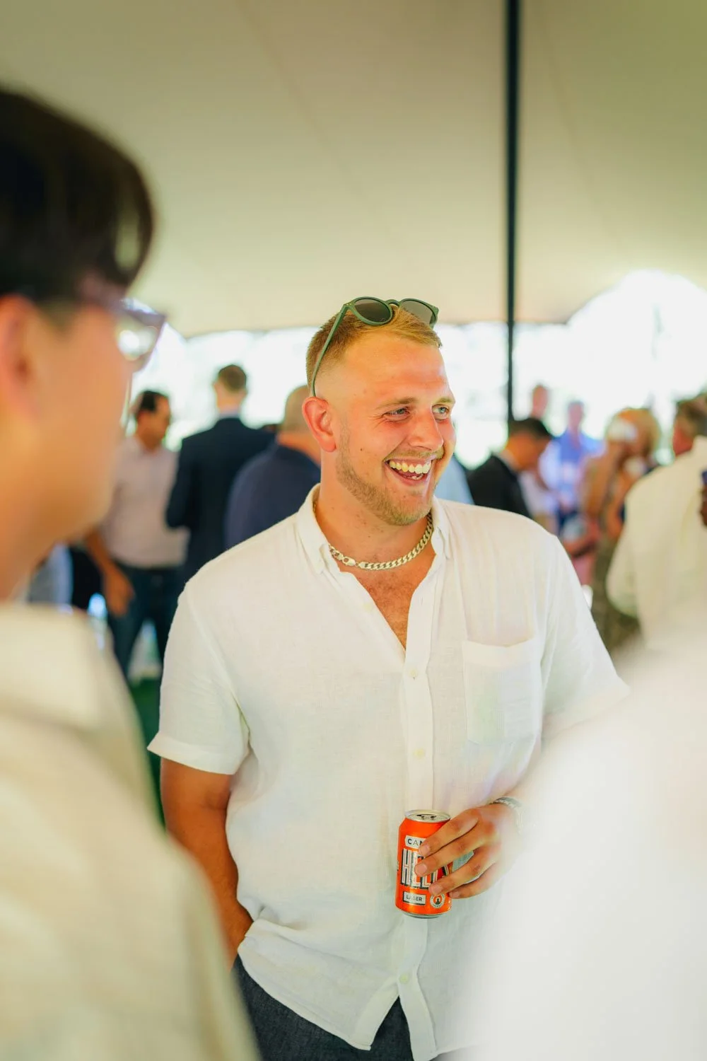 A man smiling and holding a can of soda at a social gathering under a large tent, with others in the background.