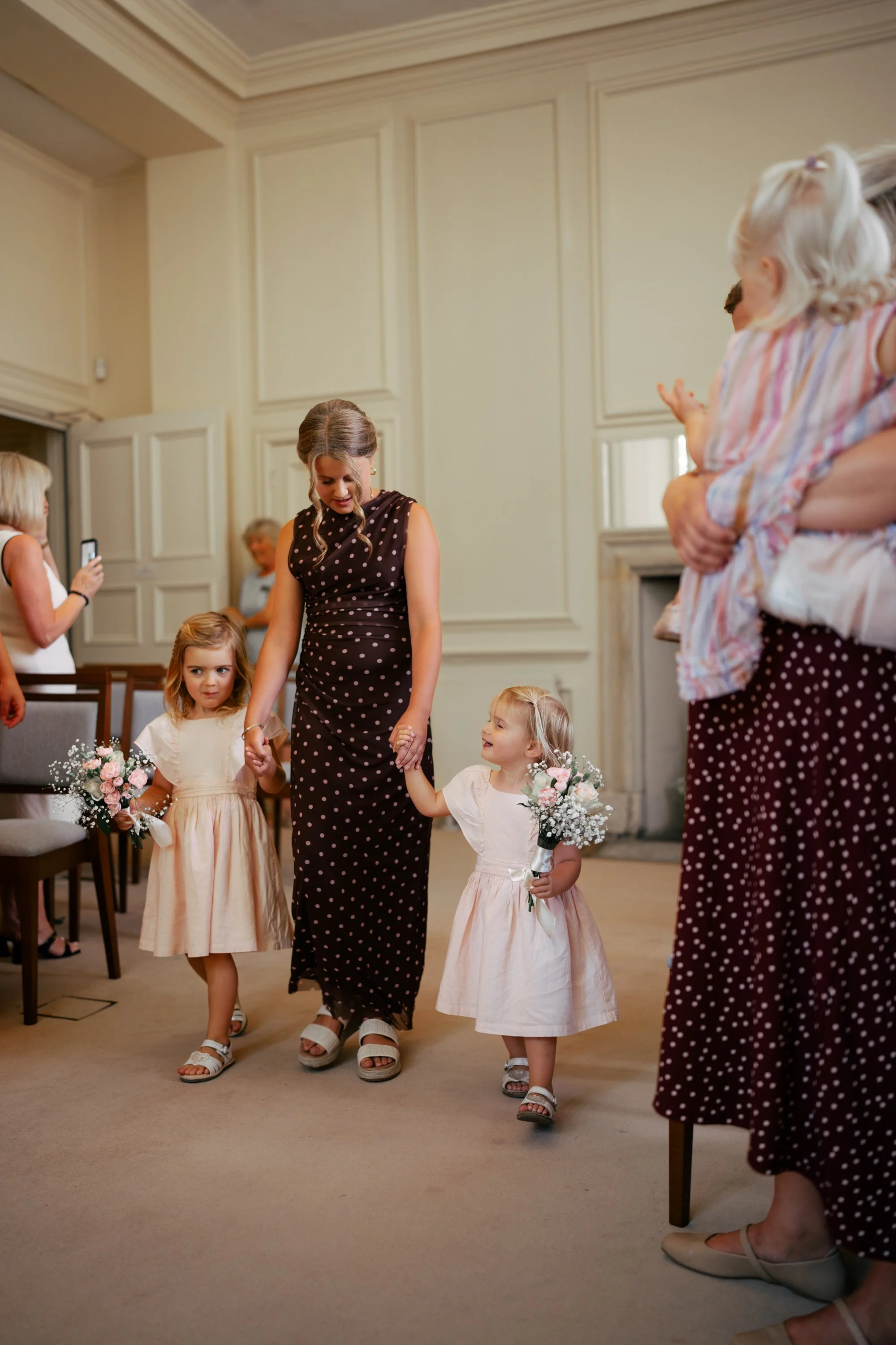 A woman dressed in a polka-dot dress is holding hands with two young girls in light-colored dresses, each holding a bouquet of flowers, in a room with beige walls and high ceilings. Other women, some taking photos, are present in the background.