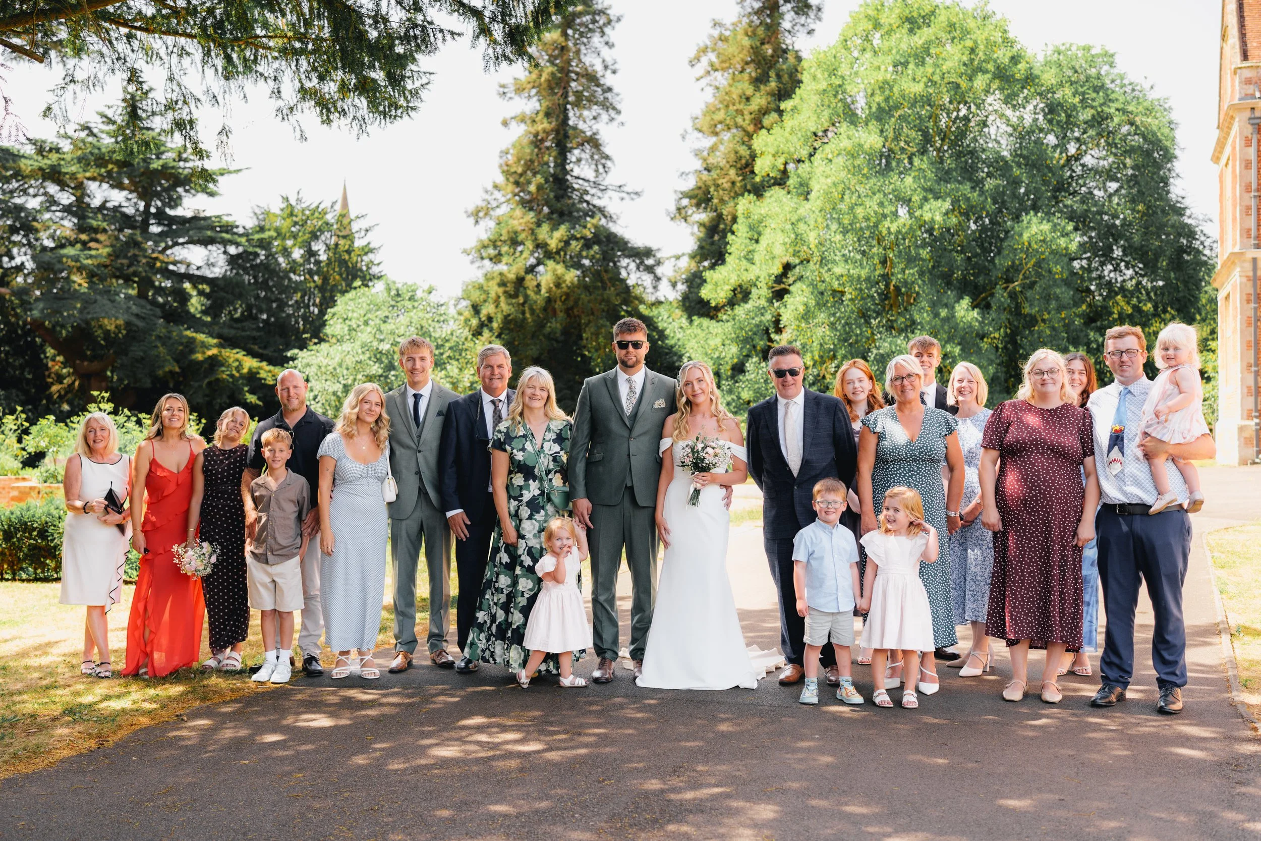 A large group of people dressed in formal and semi-formal clothing posing outdoors on a sunny day, with trees and a building in the background, at what appears to be a wedding or celebration.